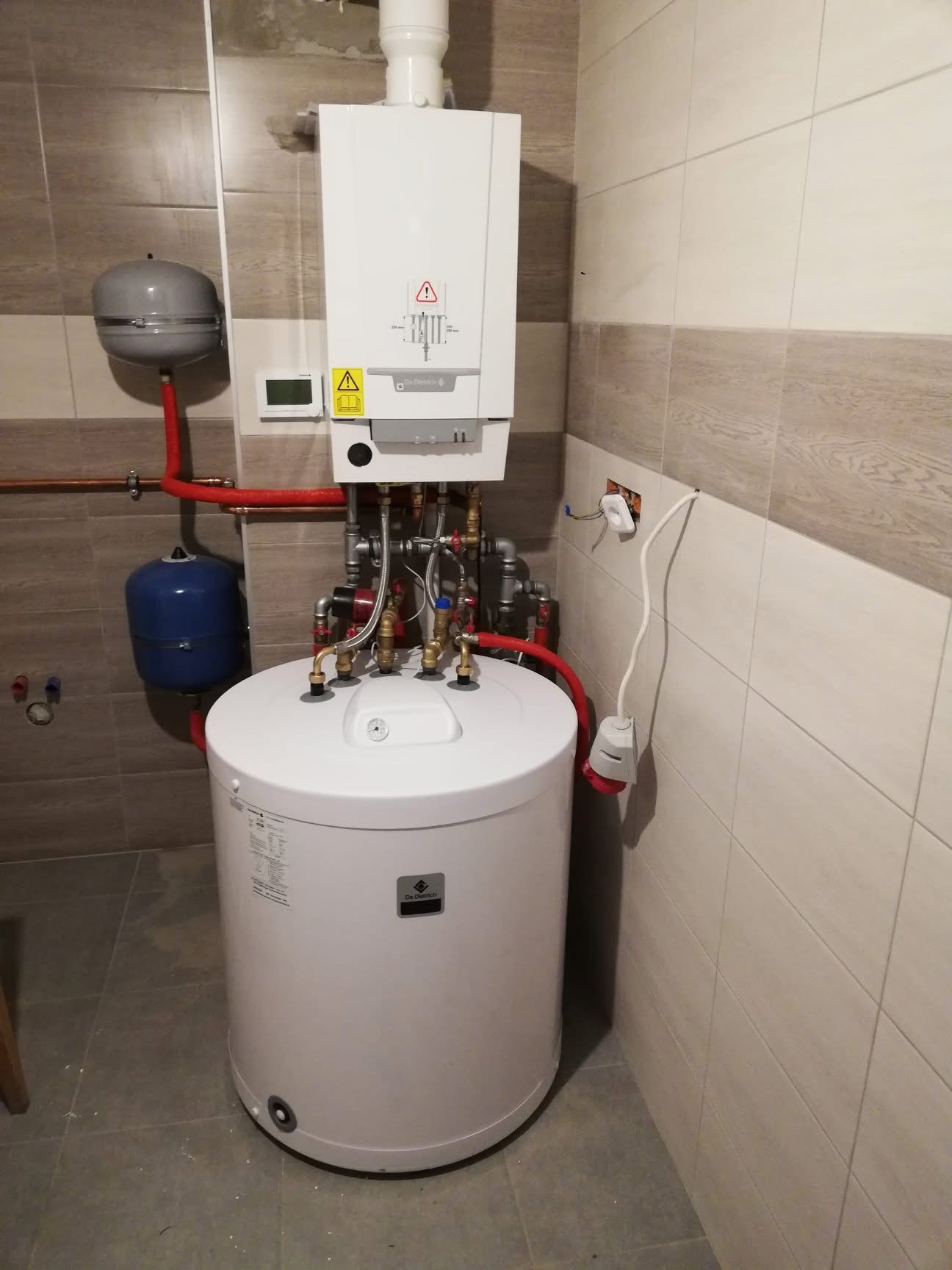 Biały, wiszący piec grzewczy marki De Dietrich z podłączonym zasobnikiem ciepłej wody, widoczne rury miedziane i zawory, całość zamontowana na tle płytek ceramicznych.