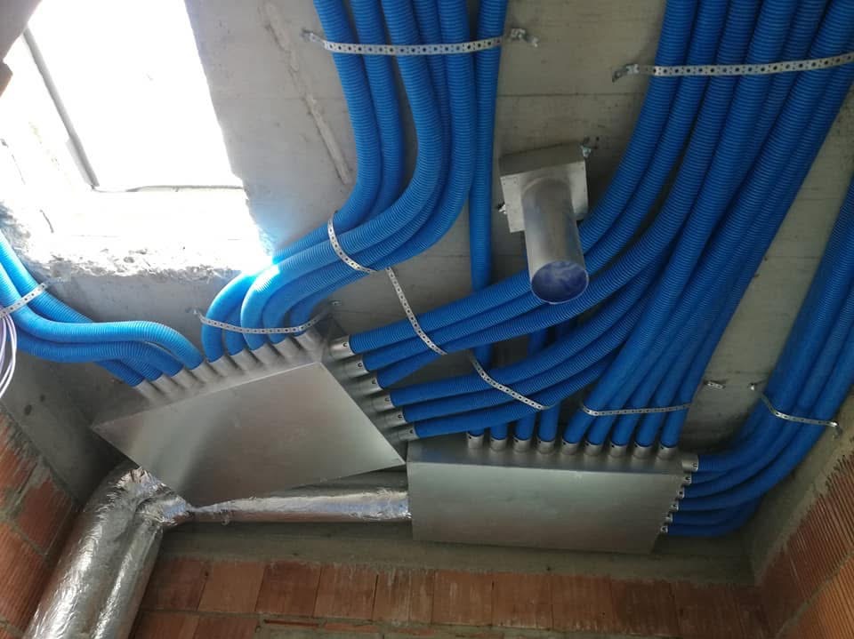 Niebieskie, karbowane rury wentylacyjne przymocowane do metalowych skrzynek rozdzielczych i sufitu betonowego za pomocą perforowanych taśm mocujących.