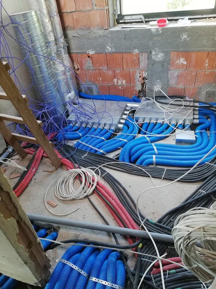 Rozłożone, niebieskie, karbowane przewody instalacyjne z metalowymi obejmami, rury wentylacyjne, czerwone i czarne kable w surowym stanie, widoczne cegły i drabina.