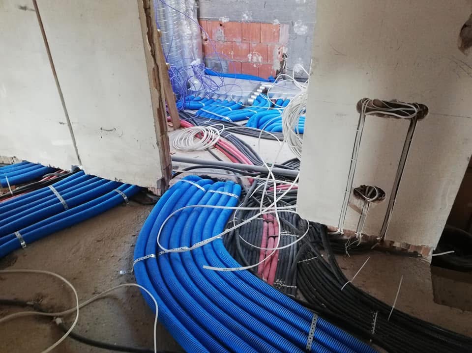 Wnętrze w trakcie prac instalacyjnych: wiązki niebieskich rur karbowanych spięte metalowymi klamrami, czarne i czerwone kable ułożone na surowej podłodze, widoczne fragmenty ścian i otworów...