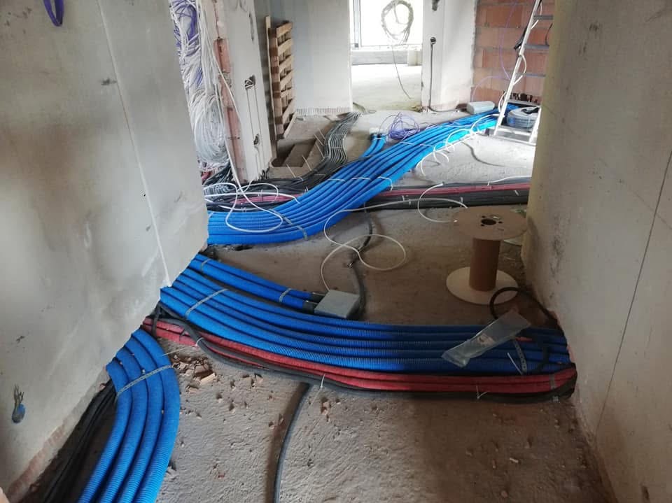 Instalacja rur karbowanych w kolorze niebieskim i czerwonym, ułożonych równolegle na podłodze w trakcie montażu w surowym stanie budynku, widoczny bęben kablowy i drabina