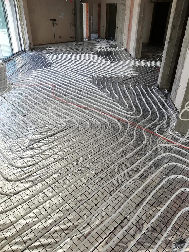 Rozłożona instalacja ogrzewania podłogowego na folii aluminiowej z siatką, widoczne białe rurki w regularnych odstępach, surowy stan wykończenia wnętrza.