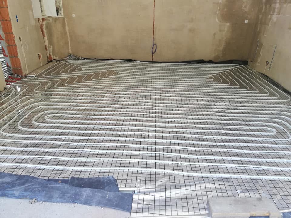 Instalacja ogrzewania podłogowego w surowym wnętrzu, rury ułożone na siatce z folią aluminiową, widoczne fragmenty betonu i instalacji elektrycznej.