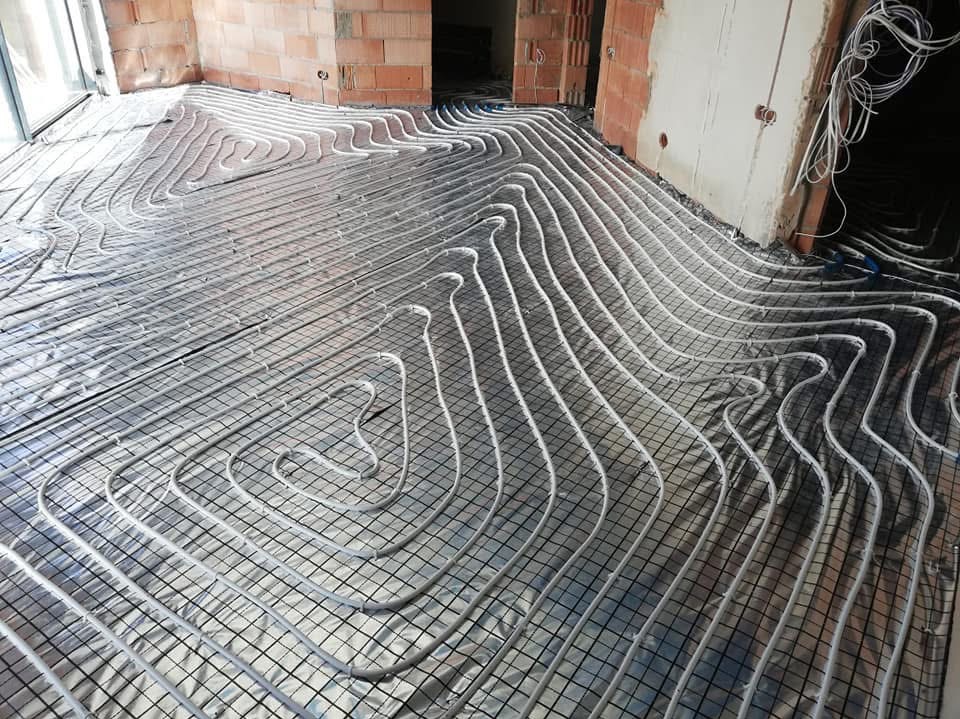 Rozłożone rury ogrzewania podłogowego na srebrnej folii izolacyjnej z siatką, surowy stan wykończenia wnętrza z widocznymi cegłami i kablami.