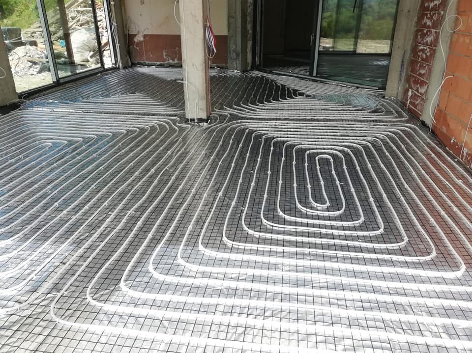 Białe rurki ogrzewania podłogowego ułożone spiralnie na siatce w surowym wnętrzu budynku w trakcie budowy, widoczne okna balkonowe i filar.