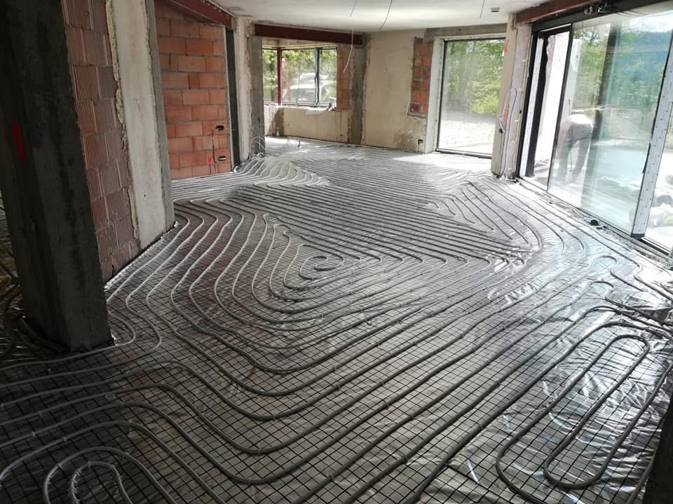 Rozłożona instalacja ogrzewania podłogowego na folii w surowym wnętrzu budynku w trakcie budowy, widoczne ceglane ściany i okna.