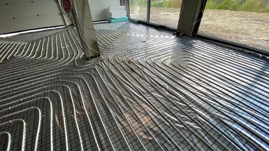 Instalacja rur grzewczych podłogowych na metalowej siatce, pokrytej folią izolacyjną, widok na surowy betonowy filar i okno z widokiem na zewnątrz.