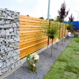 Nowoczesne ogrodzenie: gabion z kamienia połączony z drewnianymi panelami, obok trawnik z rolki, młode drzewka i wysypany kamienny żwir.