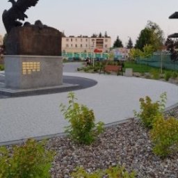 GREEN TREE Karol Kluczkowski - Uporządkowana przestrzeń miejska z pomnikiem, ścieżką wyłożoną kostką brukową, rabatami z krzewami i elementami małej architektury, takimi jak ławka, na tle budynków mieszkalnych.