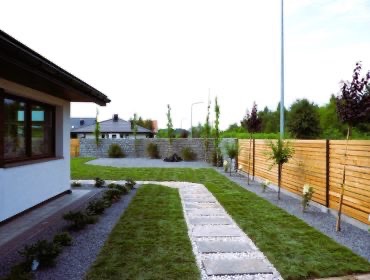 Ścieżka z płyt chodnikowych prowadząca przez świeżo założony trawnik, obok drewniany płot i młode drzewka, widok na dom z szarą elewacją i kamienny murek w tle.