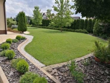 Starannie utrzymany trawnik z obrzeżami z kostki i kamienia, ozdobne krzewy i drzewa iglaste w tle, widok z perspektywy lekko z góry.