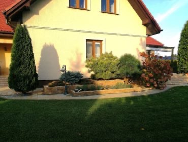 Ogród przydomowy w Łajskach z równo przystrzyżonym trawnikiem, ozdobnymi krzewami iglastymi i liściastymi posadzonymi w podwyższonych rabatach z kamienia. Widoczny fragment domu z oknem.