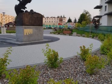 Uporządkowana przestrzeń miejska z pomnikiem, ścieżką wyłożoną kostką brukową, rabatami z krzewami i elementami małej architektury, takimi jak ławka, na tle budynków mieszkalnych.