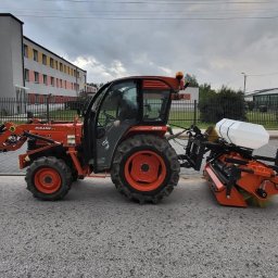 ZOSIATKA SPÓŁKA Z OGRANICZONĄ ODPOWIEDZIALNOŚCIĄ - Pomarańczowy traktor z kabiną i zamontowanym z przodu łyżką oraz z tyłu urządzeniem do pielęgnacji trawnika, stojący na asfaltowej powierzchni w pobliżu budynku.