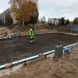 Zbrojenie pod płytę fundamentową z widocznymi pracownikami, poziomnicą laserową i izolacją obwodową, w tle budynki mieszkalne i jesienne drzewa.