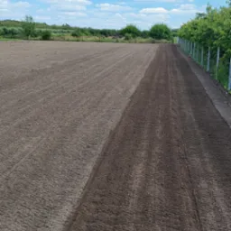 Przygotowany teren pod przyszły ogród, z widoczną granicą między ziemią wyrównaną i jeszcze nieprzetworzoną, w tle ogrodzenie i błękitne niebo z chmurami.