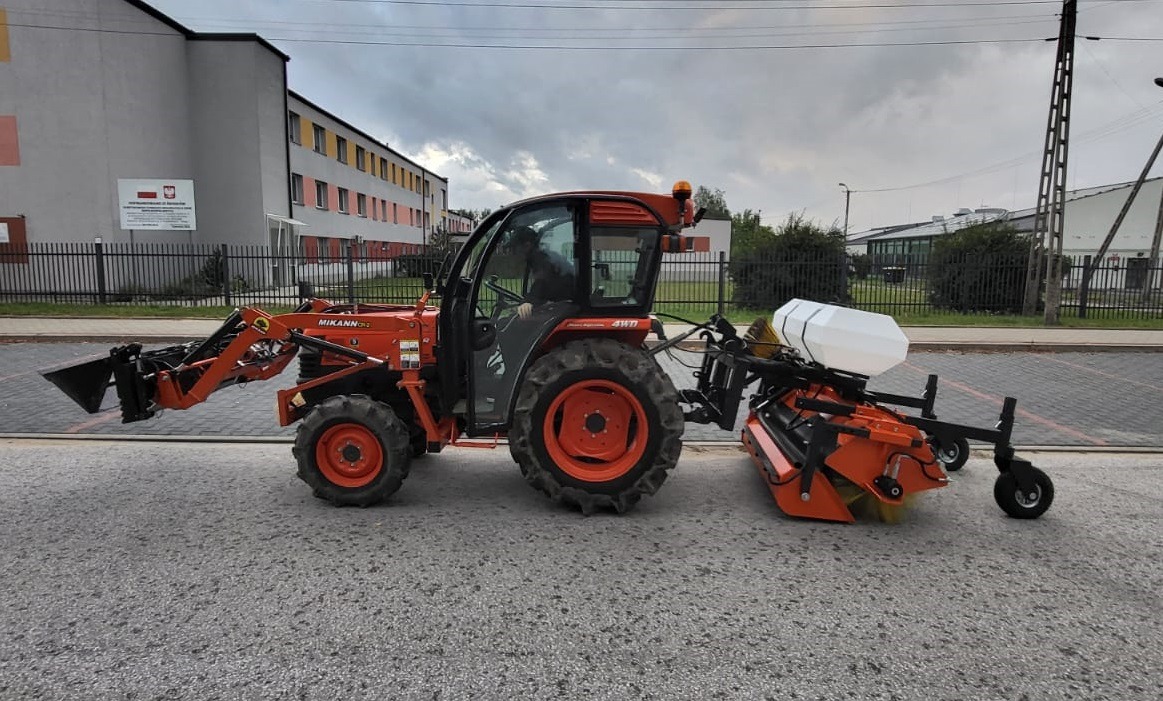 Pomarańczowy traktor z kabiną i zamontowanym z przodu łyżką oraz z tyłu urządzeniem do pielęgnacji trawnika, stojący na asfaltowej powierzchni w pobliżu budynku.