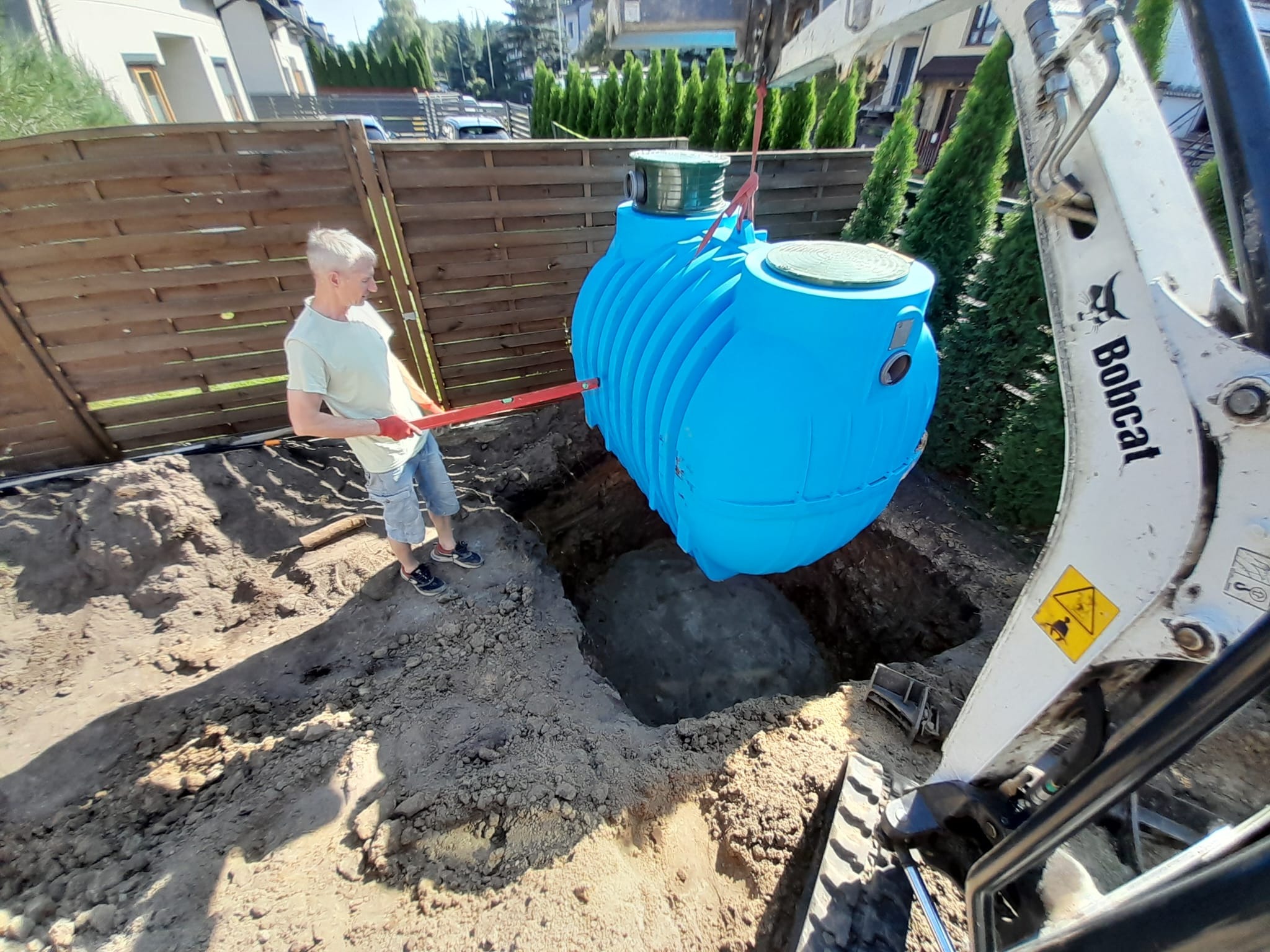 Instalacja niebieskiego zbiornika retencyjnego w wykopie przy użyciu miniładowarki Bobcat, pracownik z poziomicą kontroluje ułożenie.