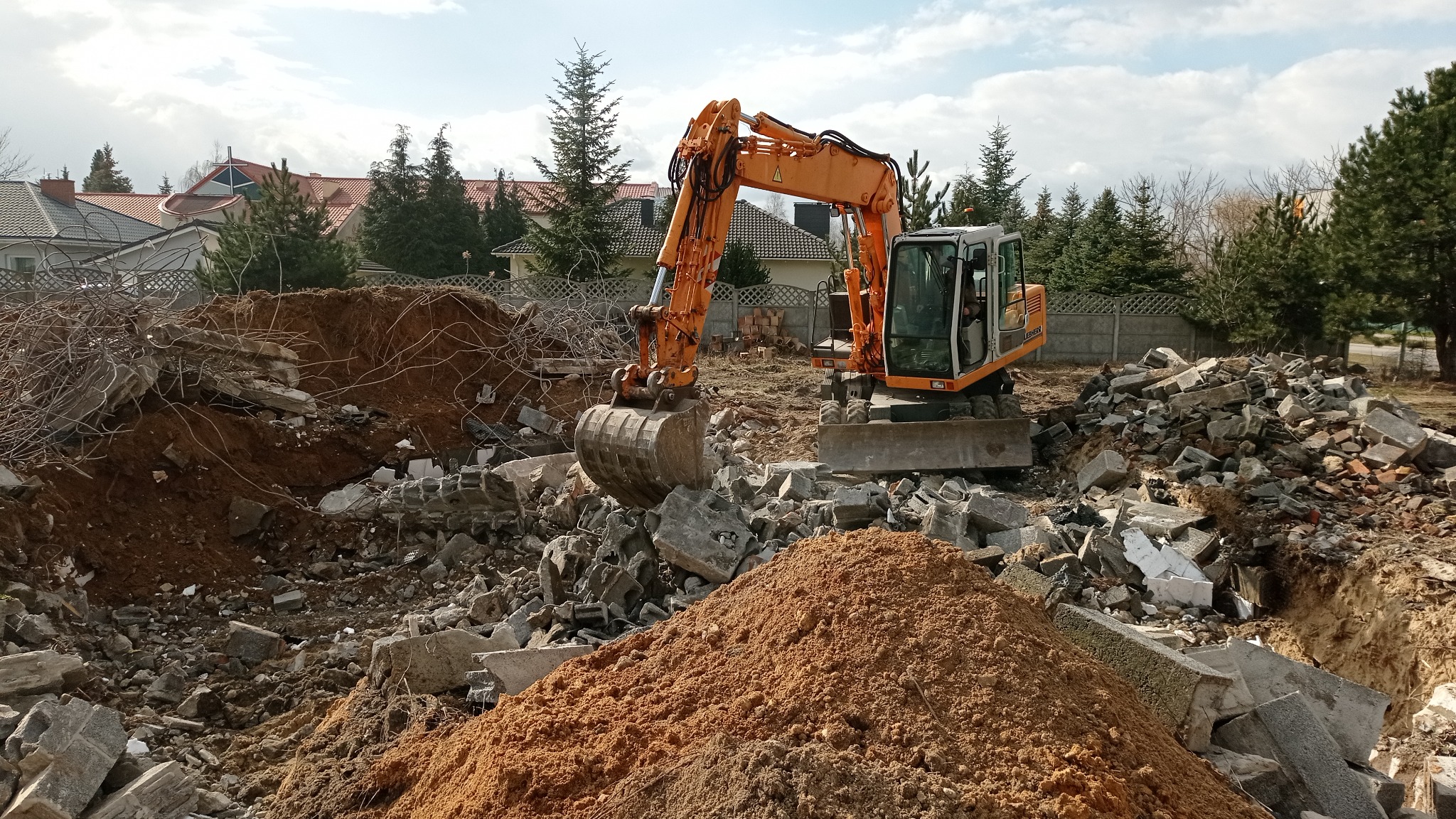 Pomarańczowa koparka Liebherr na placu budowy, w tle zwały gruzu i ziemi, widoczne budynki mieszkalne i drzewa.