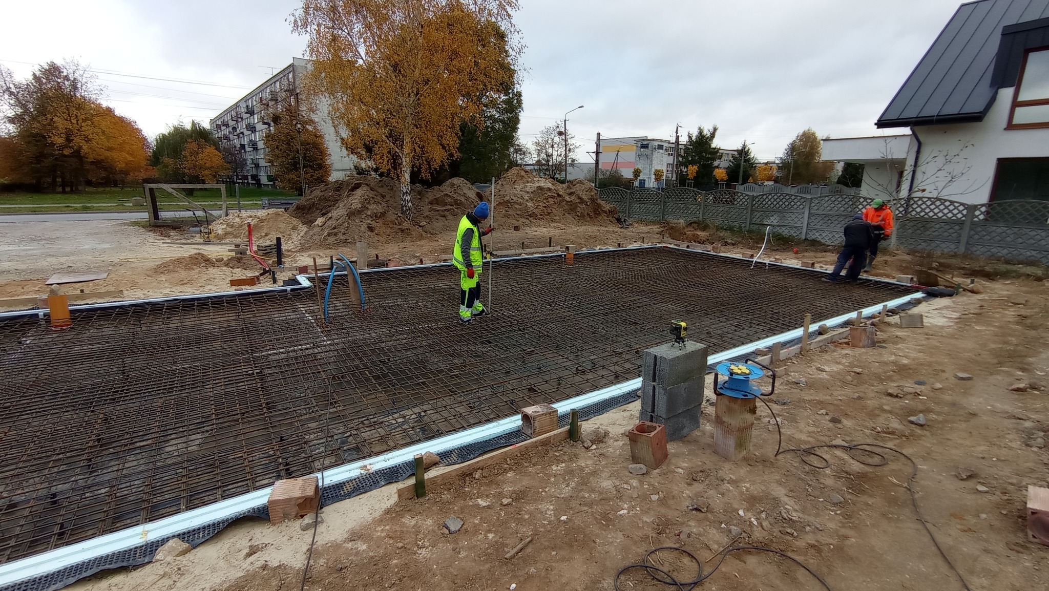 Zbrojenie pod płytę fundamentową z widocznymi pracownikami, poziomnicą laserową i izolacją obwodową, w tle budynki mieszkalne i jesienne drzewa.