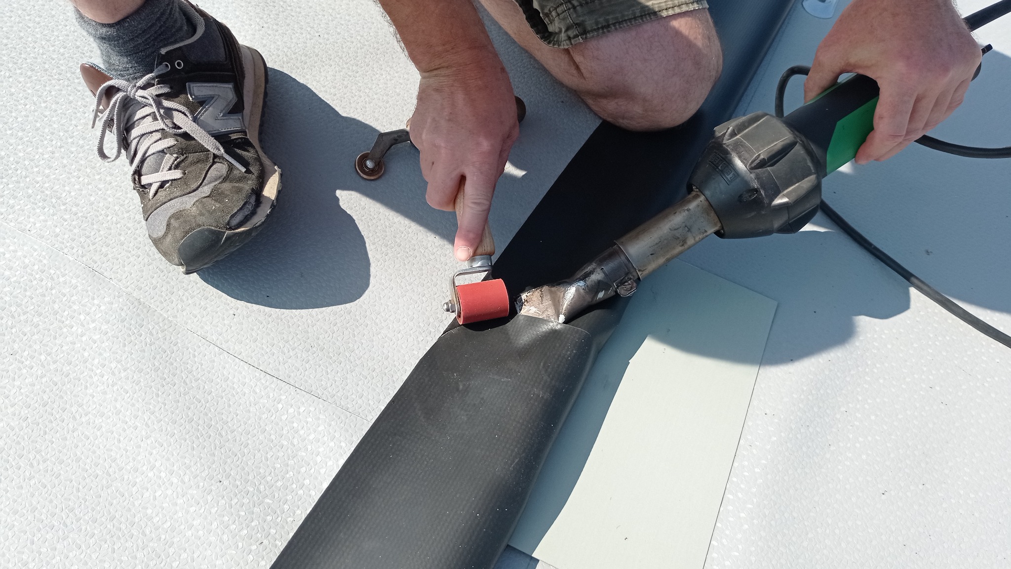 Prace dekarskie: Zgrzewanie papy termozgrzewalnej za pomocą zgrzewarki i dociskanie wałkiem, widoczny fragment obuwia roboczego.