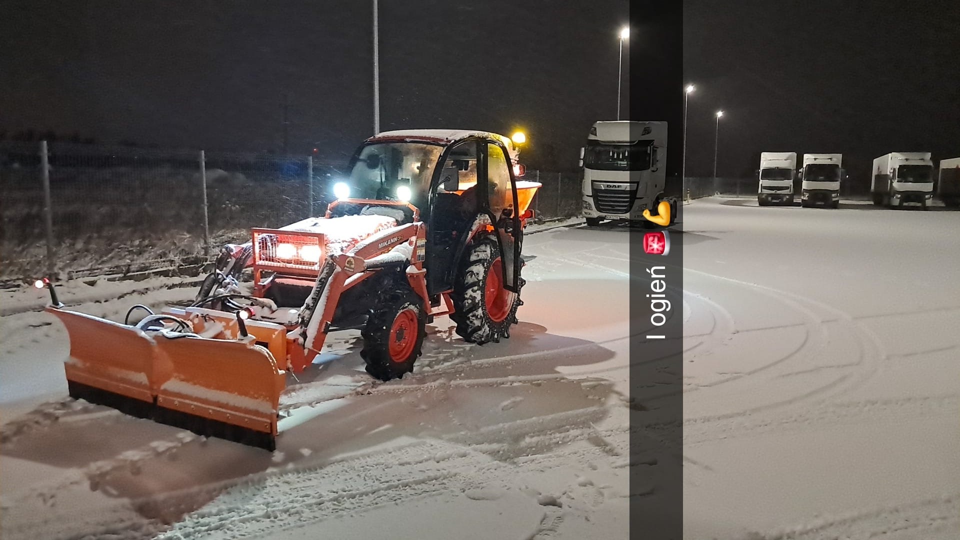 Pomarańczowy traktor z pługiem odśnieżającym, oświetlony, na zaśnieżonym parkingu z zaparkowanymi ciężarówkami w tle, nocna sceneria.