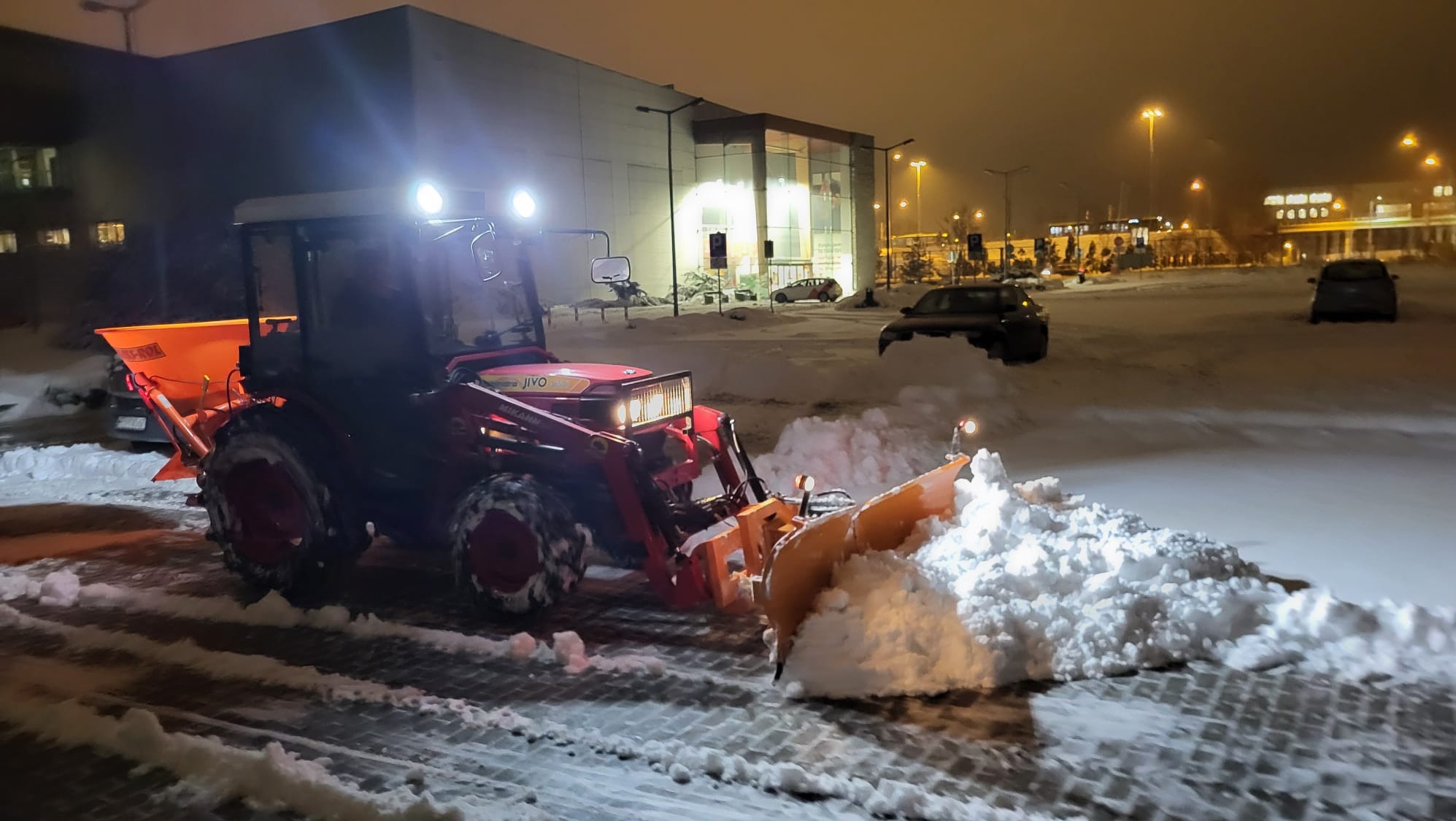 Czerwony traktor z pługiem odśnieża parking przed budynkiem w nocnych warunkach, widoczne ślady opon na śniegu i zaparkowane samochody.