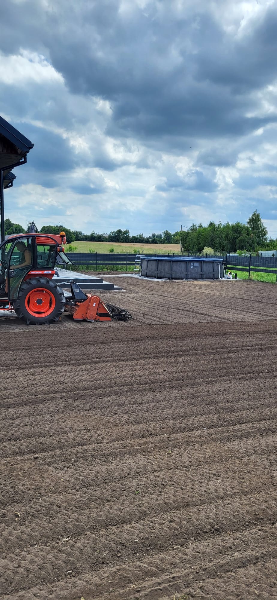 Czerwony traktor z glebogryzarką przygotowuje ziemię pod trawnik, widoczny basen ogrodowy i fragment domu.