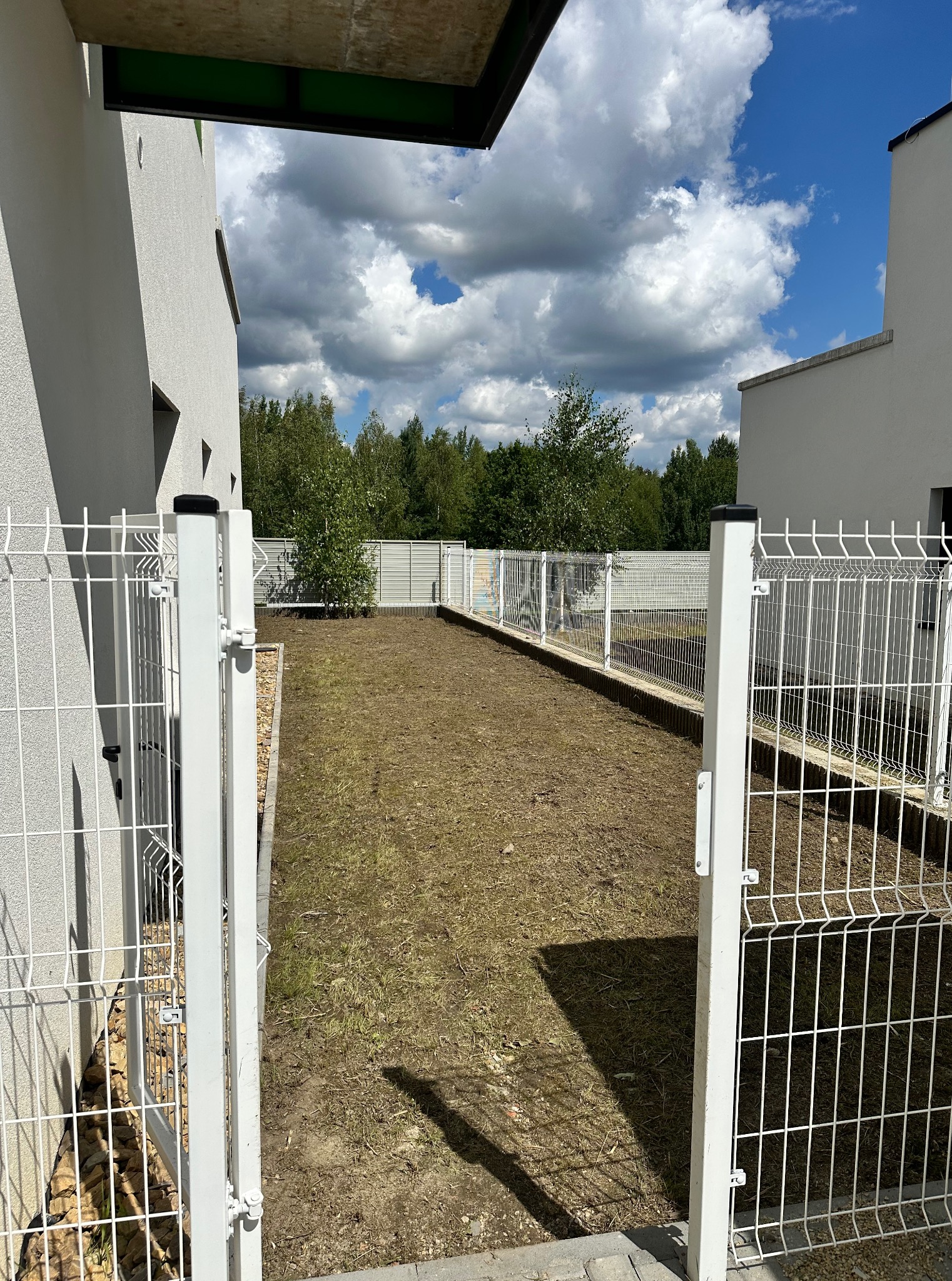 Widok na trawnik otoczony białym ogrodzeniem panelowym, z otwartą furtką na pierwszym planie. W tle drzewa i błękitne niebo z chmurami.