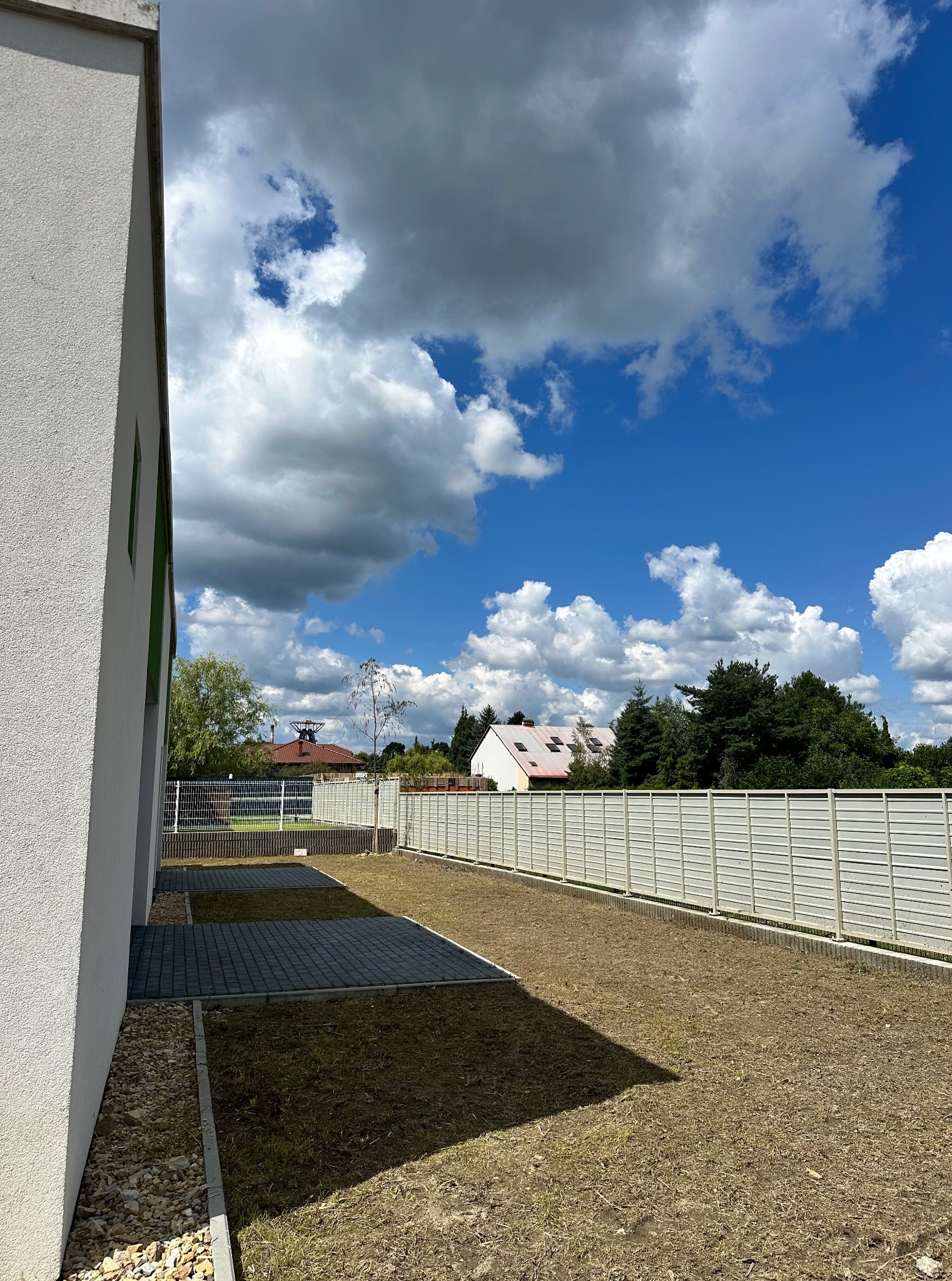 Ogród z świeżo posianą trawą przy budynku i ogrodzeniu panelowym w słoneczny dzień. Widoczne chmury na błękitnym niebie.