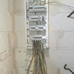 VOLTAGE SZYMON KACZYKOWSKI - Szafa rozdzielcza w trakcie montażu, z licznymi przewodami wychodzącymi dołem, zamontowana na surowej ścianie z betonu komórkowego.