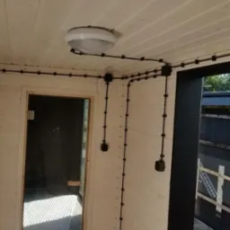 Wnętrze sauny z widoczną instalacją elektryczną natynkową w czarnych peszlach, lampa sufitowa w centrum kadru, drzwi szklane do sauny po lewej stronie.