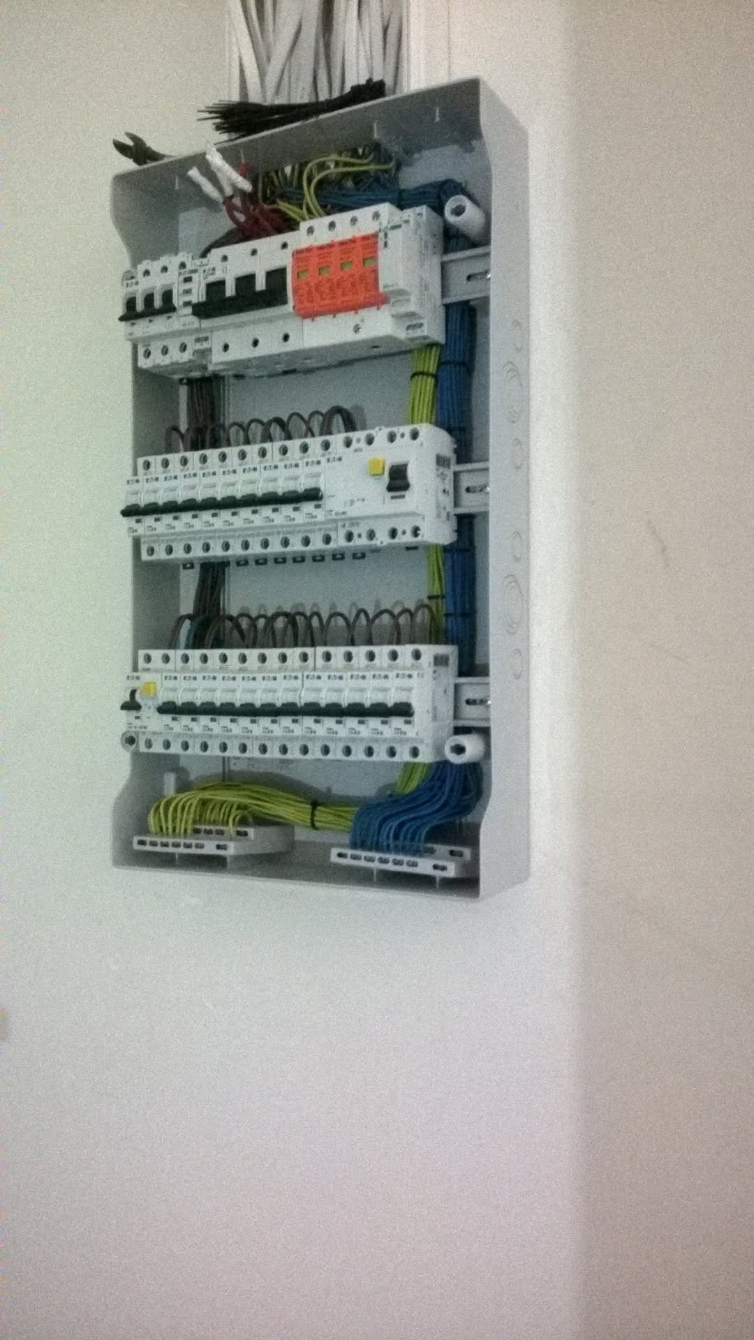 Otwarta skrzynka rozdzielcza z widocznymi bezpiecznikami, przewodami w kolorach żółto-zielonym i niebieskim, oraz narzędziami na górze, zamontowana na białej ścianie.