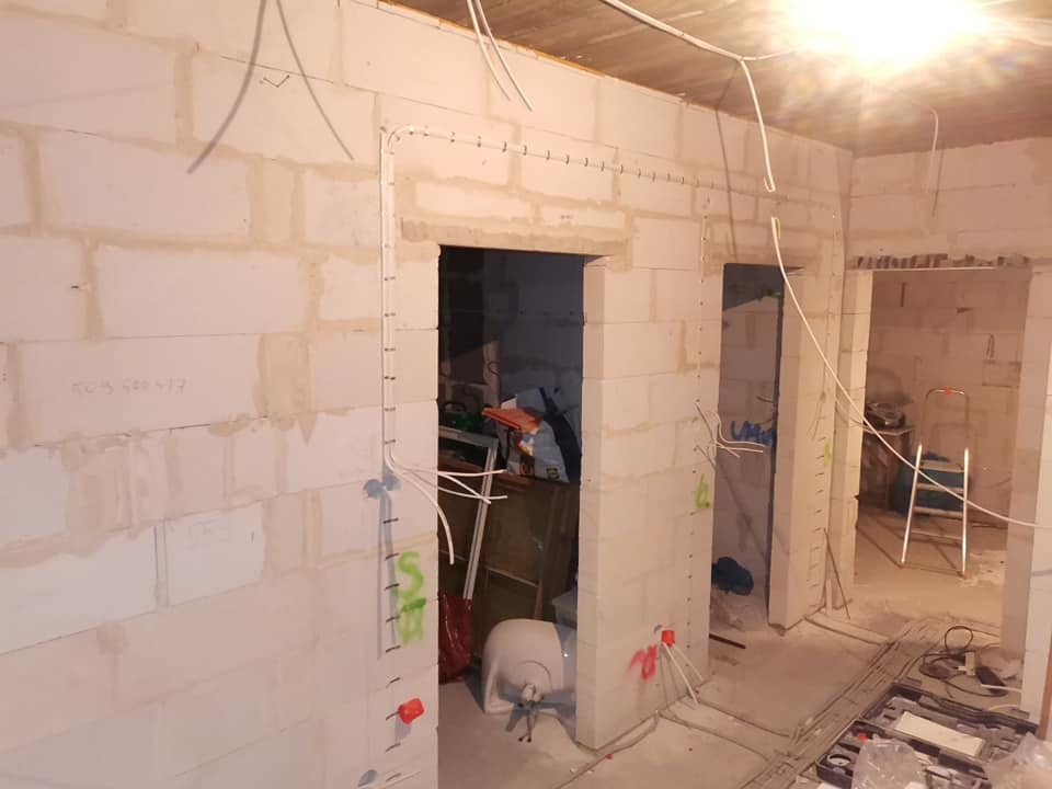 Wnętrze w trakcie prac instalacyjnych: białe ściany z betonu komórkowego, widoczne przewody elektryczne prowadzone po ścianach, oznaczenia na ścianach, otwarte przejścia między pomieszczeniami...