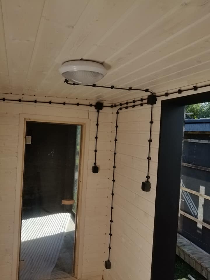 Wnętrze sauny z widoczną instalacją elektryczną natynkową w czarnych peszlach, lampa sufitowa w centrum kadru, drzwi szklane do sauny po lewej stronie.