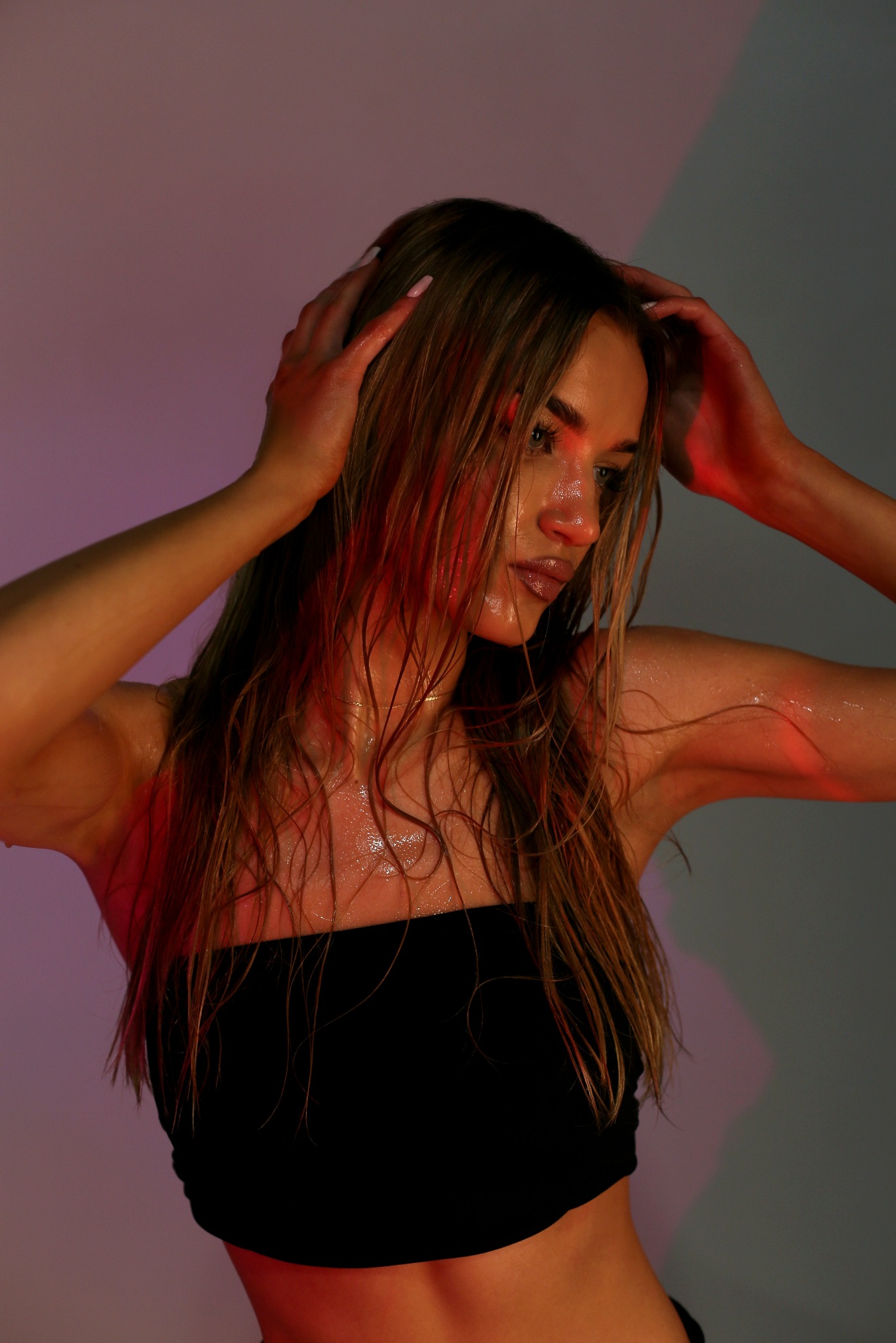 Portret kobiety z mokrymi włosami, pozującej w czarnym topie na tle z czerwonym i szarym oświetleniem.