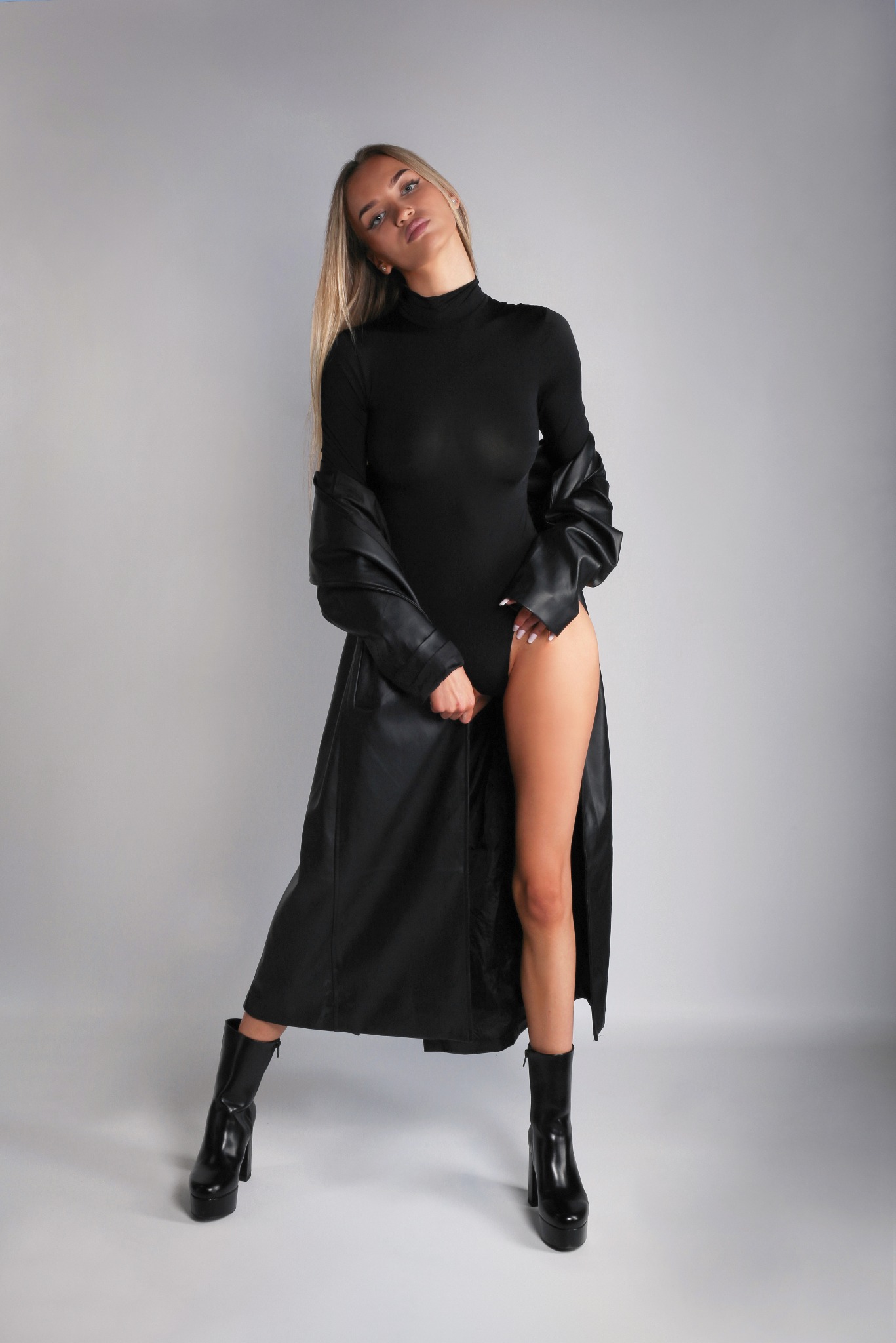 Kobieta w czarnym body, płaszczu z imitacji skóry i botkach na platformie, pozująca na jasnoszarym tle w studio.