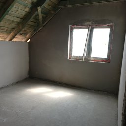 Ł&A Tynki Maszynowe - Surowe wnętrze poddasza z oknem dachowym i świeżo nałożonym tynkiem na ścianach, widoczne drewniane belki stropowe.