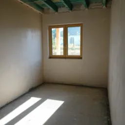 Wnętrze pokoju po nałożeniu gładzi gipsowej na ścianach, z widocznym oknem z drewnianą ramą i surowym betonowym podłożem, na którym widać smugi światła słonecznego.