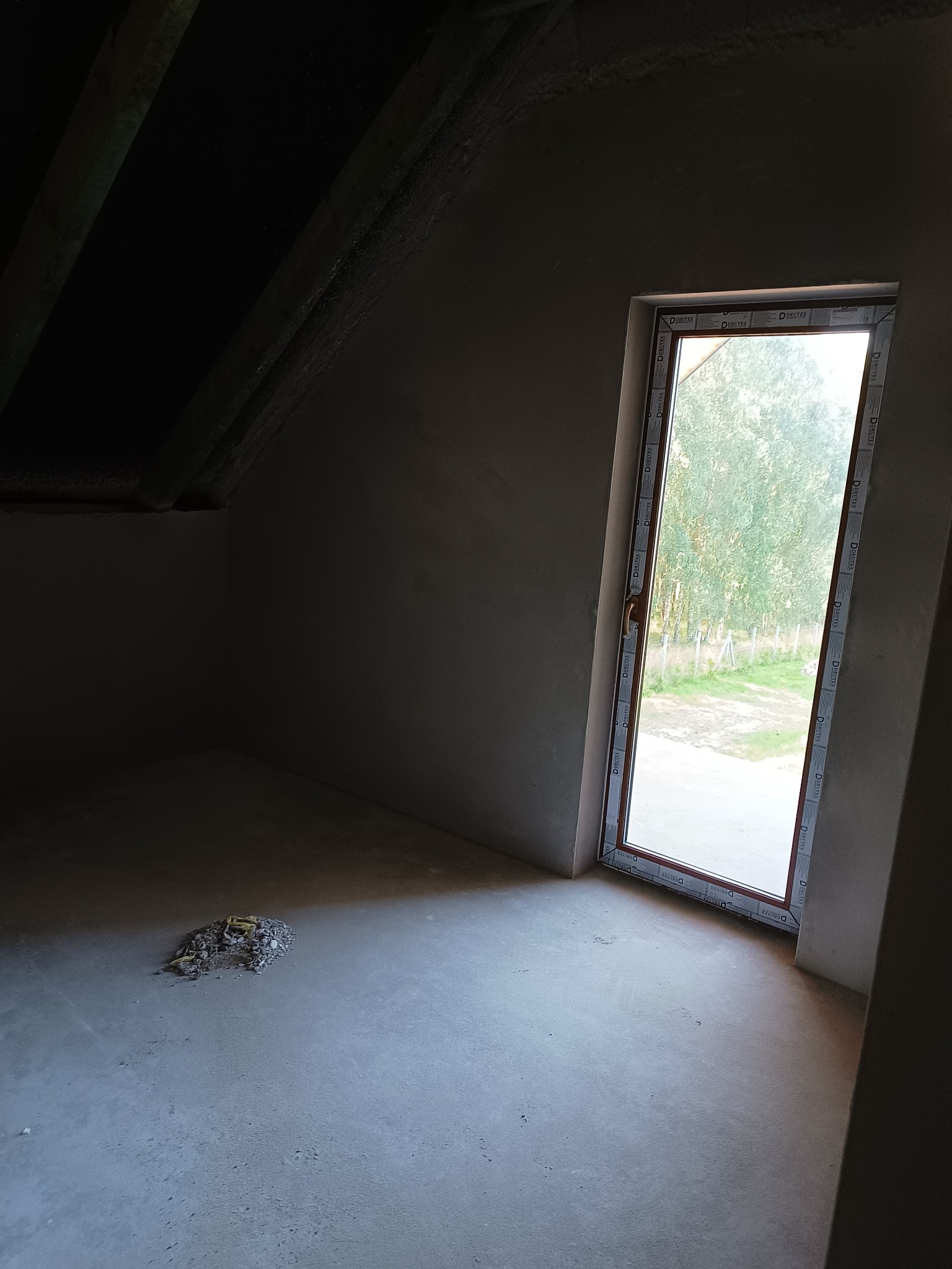 Surowe wnętrze poddasza z oknem dachowym, ściany po nałożeniu tynku, na podłodze resztki materiałów budowlanych.