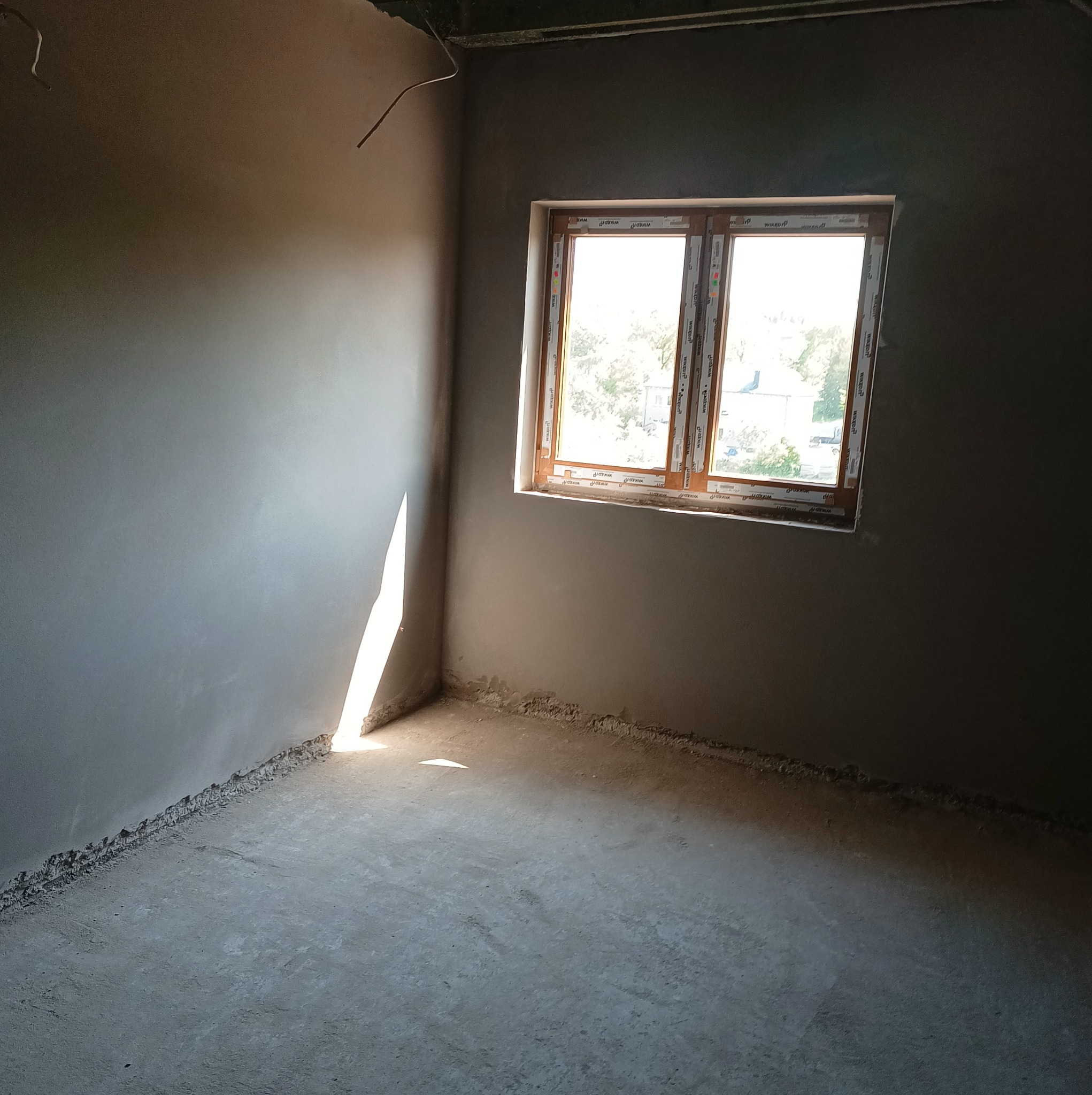 Wnętrze po tynkowaniu maszynowym w Choczewie, widoczne świeżo otynkowane ściany i podłoga, z oknem w drewnianej ramie, przez które wpada światło słoneczne.