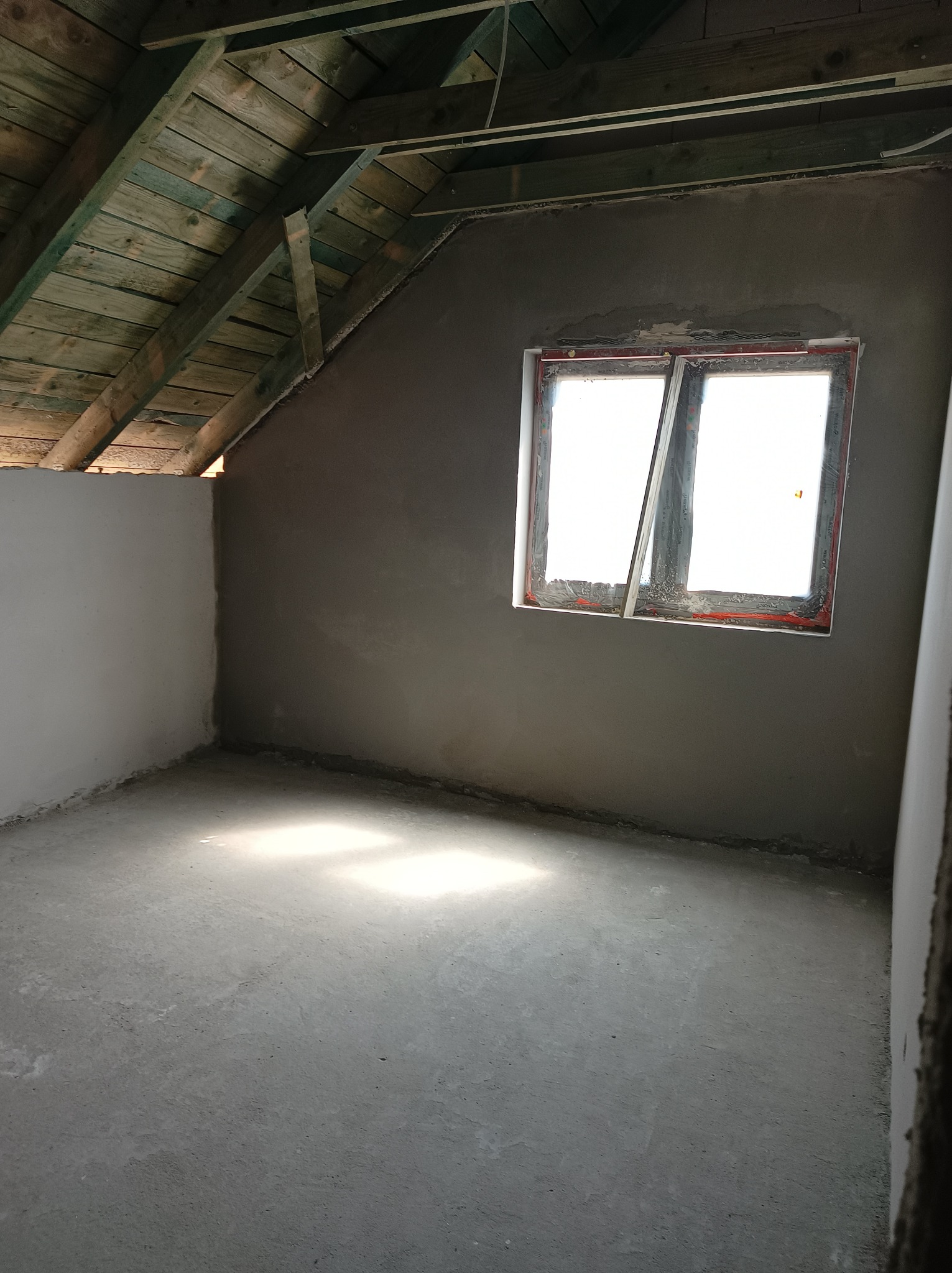 Surowe wnętrze poddasza z oknem dachowym i świeżo nałożonym tynkiem na ścianach, widoczne drewniane belki stropowe.