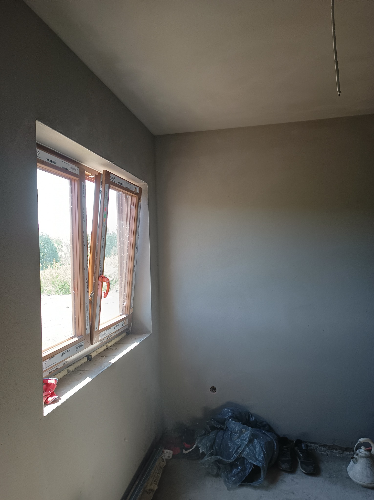 Wnętrze pokoju po nałożeniu gładzi gipsowej, widoczne okno z częściowo otwartymi skrzydłami, zabezpieczone folią podłogi i narzędzia malarskie.
