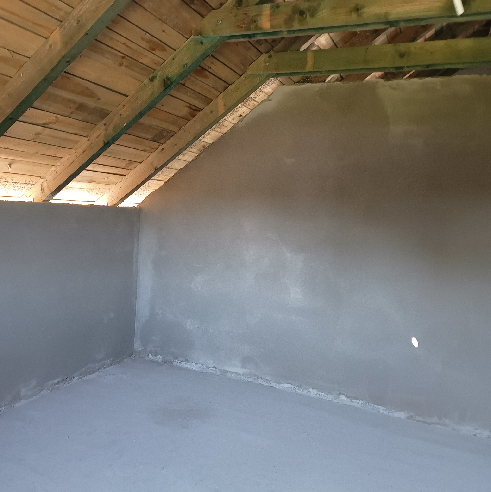 Wnętrze poddasza po tynkowaniu maszynowym, widoczne drewniane elementy konstrukcji dachu i gładka, szara powierzchnia ścian oraz podłogi, gotowa do dalszych prac wykończeniowych.