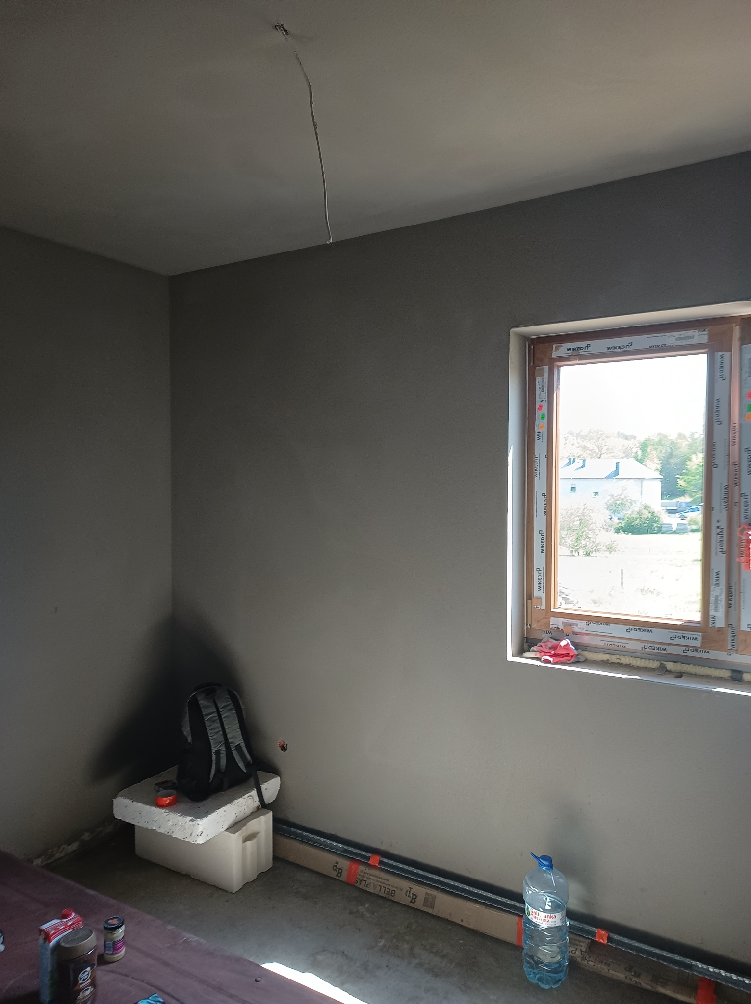 Wnętrze pokoju po nałożeniu szarego tynku maszynowego, widoczne okno z białymi ramami, wystający kabel z sufitu, plecak oparty o ścianę na styropianowym bloku, butelka wody mineralnej i profile...