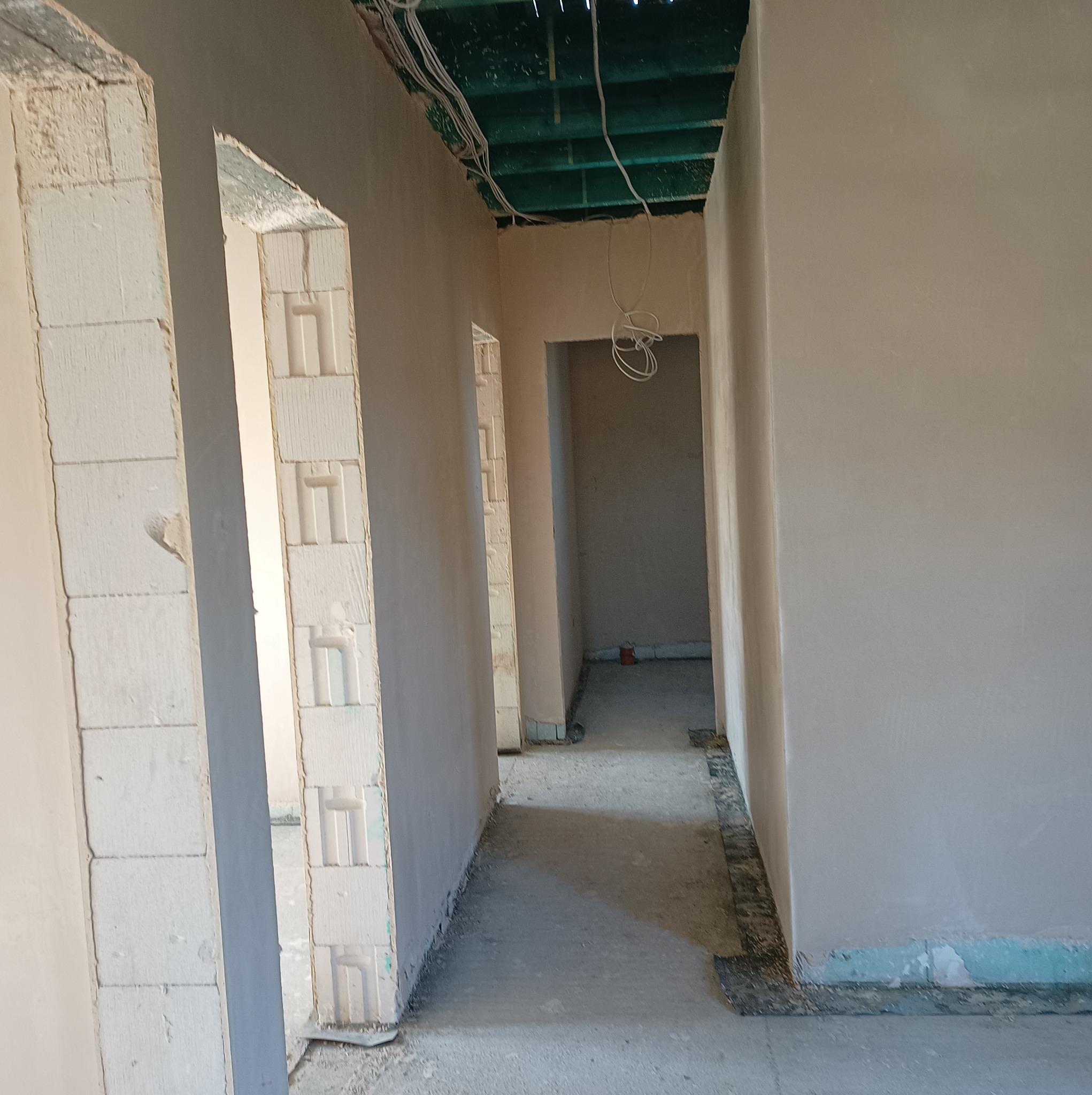 Wnętrze w trakcie prac wykończeniowych, widoczne ściany po nałożeniu tynku, surowy strop z instalacją elektryczną i niezabudowane otwory drzwiowe z bloczków.