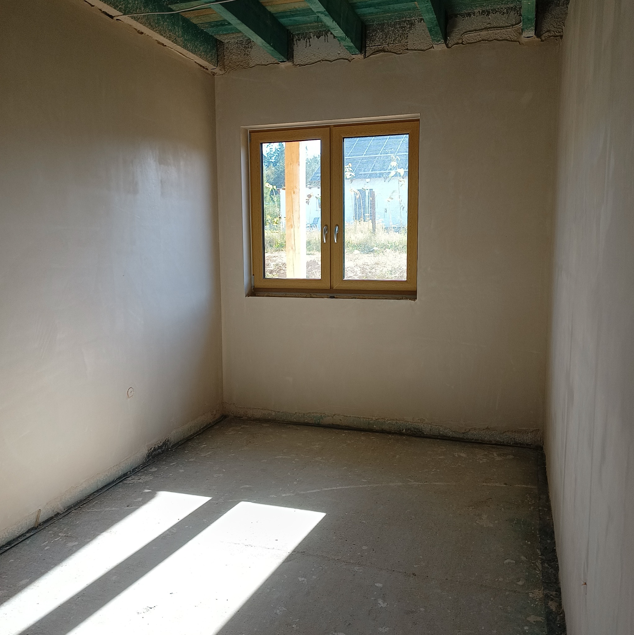 Wnętrze pokoju po nałożeniu gładzi gipsowej na ścianach, z widocznym oknem z drewnianą ramą i surowym betonowym podłożem, na którym widać smugi światła słonecznego.