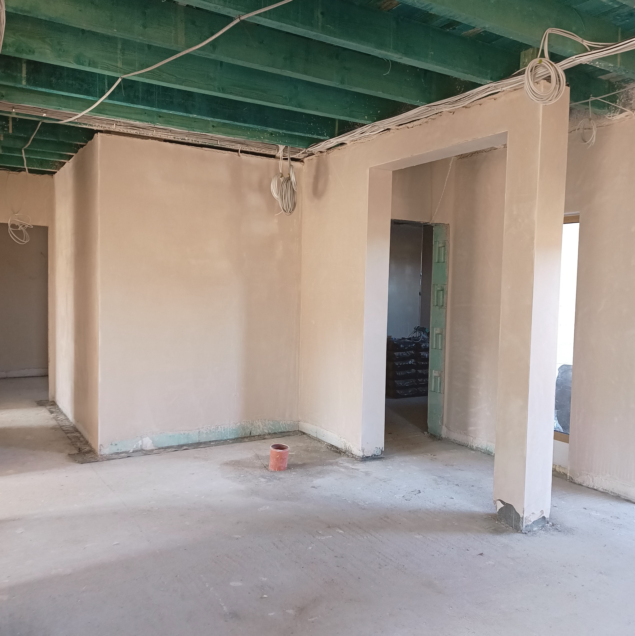 Surowe wnętrze po nałożeniu gładzi gipsowej, widoczne przewody elektryczne pod zielonym stropem, przygotowane otwory drzwiowe i okienne, na podłodze mały pomarańczowy kubełek.