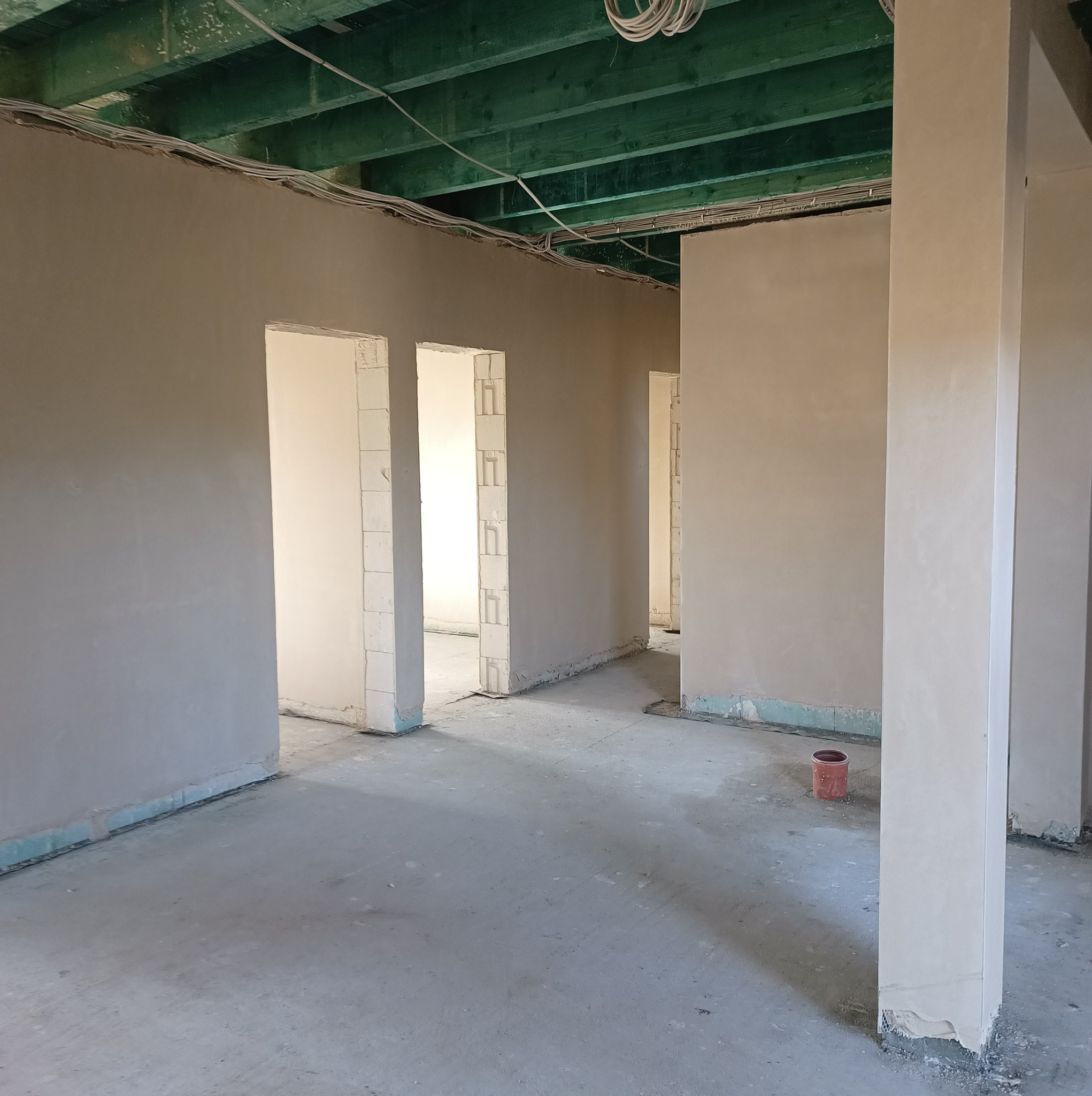 Wnętrze w stanie surowym z nałożonymi tynkami na ścianach, widoczne otwory drzwiowe z surowymi framugami i fragmentem instalacji elektrycznej pod sufitem. Na podłodze stoi czerwone wiadro.