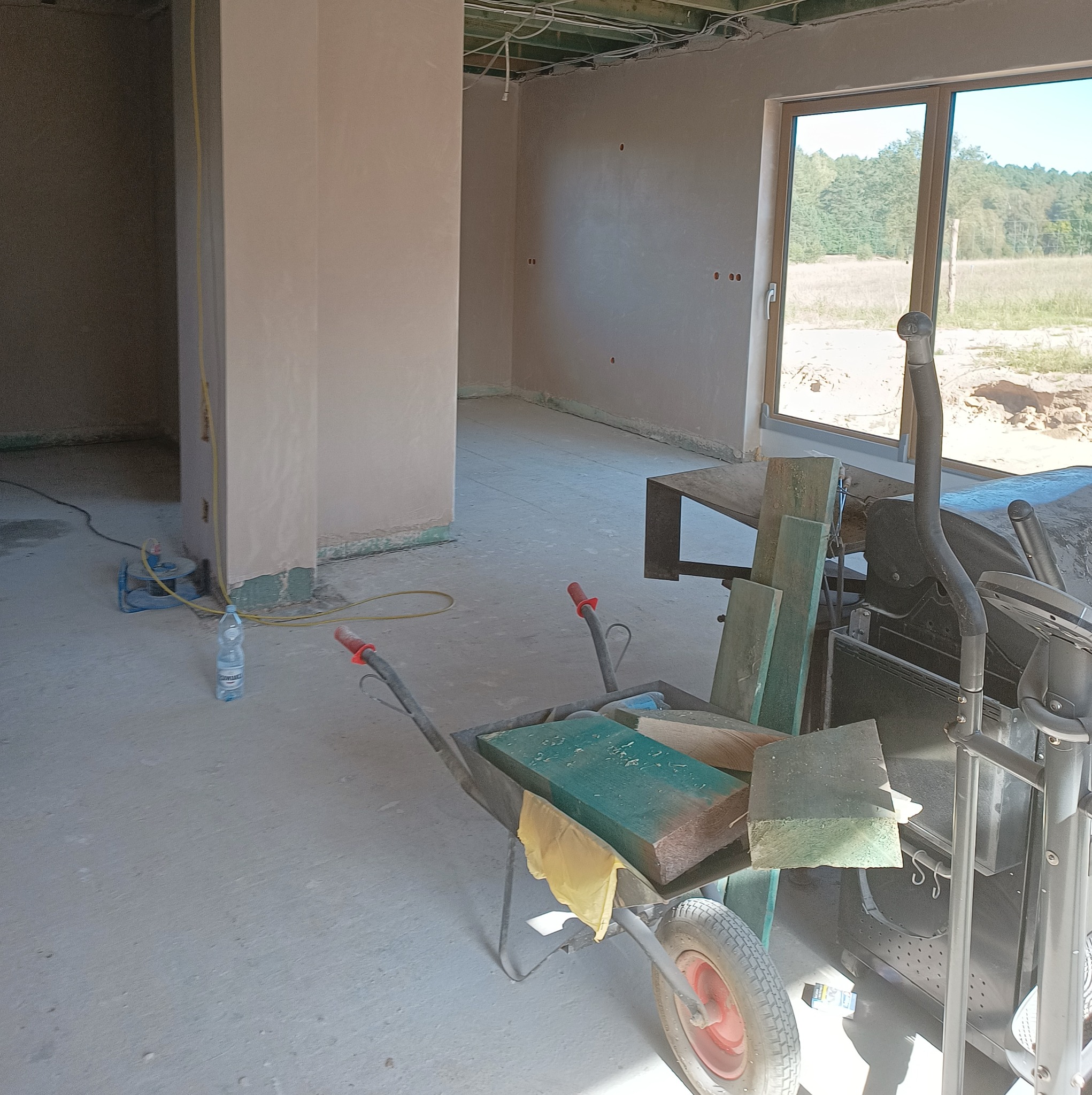 Surowe wnętrze w trakcie prac wykończeniowych: świeżo otynkowane ściany, widoczne instalacje pod sufitem, wózek budowlany z fragmentami drewna i butelka wody mineralnej na podłodze.